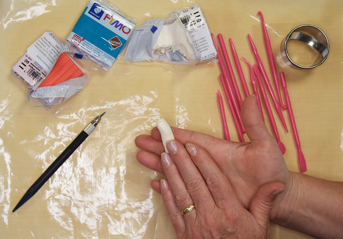 Žena rozpracováva Fimo hmotu slonovinovej farby. Na stole sú položené ostatné Fimo hmoty a modelovacie nástroje so sklapelom.